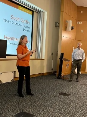 Heather Hanna, Executive Director of Enterprise Business IT Solutions (eBITS), in a burnt orange UT Works polo shirt and black pants and Scott Griffin, Interim Director of Facilities Services, in a gray business shirt and brown slacks, stand in front of an auditorium before a presentation slide listing their names and titles. 