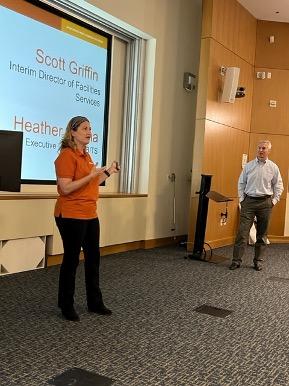 Heather Hanna, Executive Director of Enterprise Business IT Solutions (eBITS), in a burnt orange UT Works polo shirt and black pants and Scott Griffin, Interim Director of Facilities Services, in a gray business shirt and brown slacks, stand in front of an auditorium before a presentation slide listing their names and titles. 